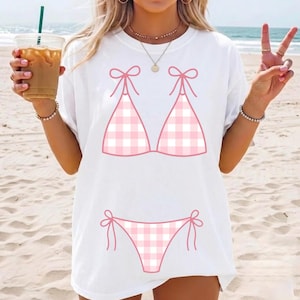 May include: White t-shirt with a pink and white gingham bikini graphic. The bikini top has bow details, and the bottoms also feature bows. The design is centrally placed on the front of the tee.