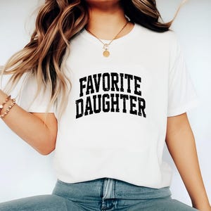Puede incluir: Camiseta blanca con la frase "FAVORITE DAUGHTER" en fuente negra desgastada. La camiseta es de cuello redondo y parece estar hecha de un material suave. La camiseta la lleva una persona.
