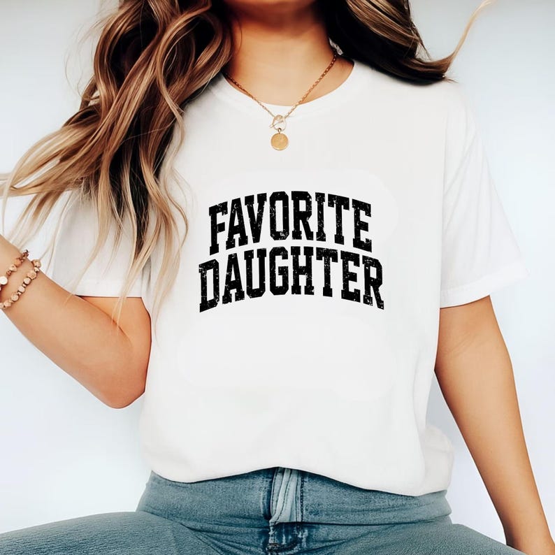 Puede incluir: Camiseta blanca con la frase "FAVORITE DAUGHTER" en fuente negra desgastada. La camiseta es de cuello redondo y parece estar hecha de un material suave. La camiseta la lleva una persona.