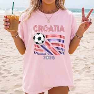 May include: A light pink short-sleeve t-shirt featuring the text CROATIA in red, a black and white soccer ball graphic, and a stylized flag design composed of red and blue horizontal lines, with the number 2026 printed in blue at the bottom.