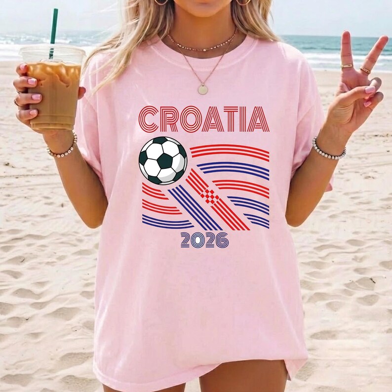 May include: A light pink short-sleeve t-shirt featuring the text CROATIA in red, a black and white soccer ball graphic, and a stylized flag design composed of red and blue horizontal lines, with the number 2026 printed in blue at the bottom.