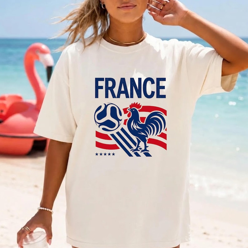 Puede incluir: Una camiseta de manga corta color hueso con la palabra FRANCE en letras may&uacute;sculas azules sobre un gr&aacute;fico de un gallo azul, un bal&oacute;n de f&uacute;tbol y rayas rojas y blancas. Cinco peque&ntilde;as estrellas azules se sit&uacute;an bajo el dise&ntilde;o gr&aacute;fico.