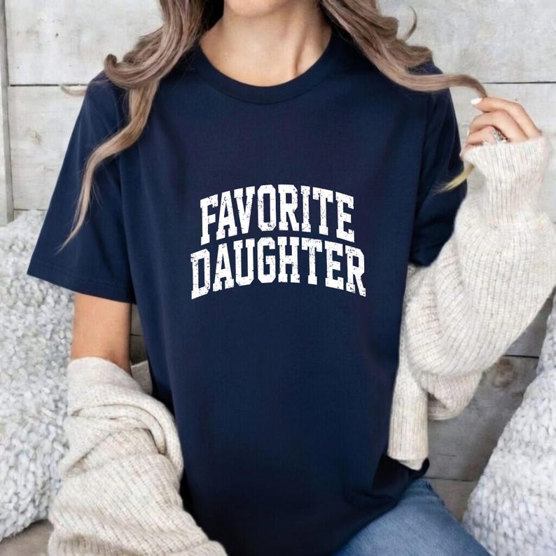 Puede incluir: Camiseta azul marino con las palabras "FAVORITE DAUGHTER" impresas en letras blancas. La camiseta es de manga corta y cuello redondo.