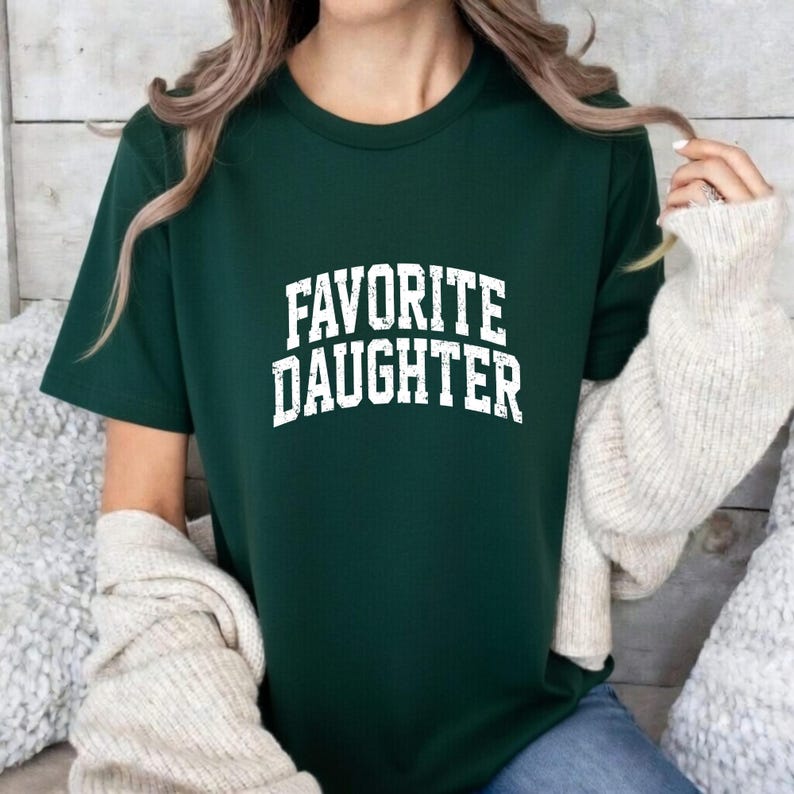 Puede incluir: Una camiseta verde oscuro con la frase "FAVORITE DAUGHTER" en letras blancas. La camiseta tiene mangas cortas y cuello redondo. El texto tiene un aspecto ligeramente desgastado.