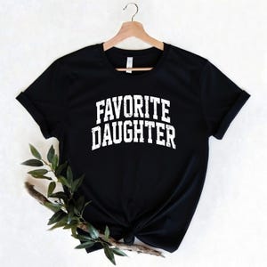 Puede incluir: Camiseta negra con el texto "FAVORITE DAUGHTER" en letras blancas. La camiseta est&aacute; colgada de una percha de madera. Una ramita de vegetaci&oacute;n se coloca delante de la camiseta.