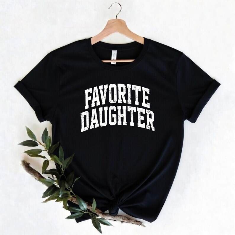 Puede incluir: Camiseta negra con el texto "FAVORITE DAUGHTER" en letras blancas. La camiseta est&aacute; colgada de una percha de madera. Una ramita de vegetaci&oacute;n se coloca delante de la camiseta.