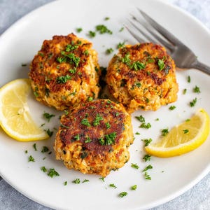 May include: Three golden-brown crab cakes on a white plate, garnished with fresh parsley. Two lemon wedges are placed on the plate. A silver fork is visible. The crab cakes have a crispy exterior and a soft interior.