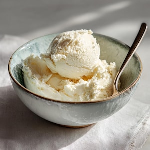 May include: A serving of vanilla ice cream in a pale blue bowl with a spoon. The ice cream is a creamy white colour and has a soft texture. The bowl has a textured rim and sits on a white cloth.