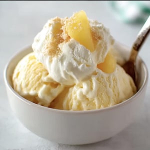 May include: A white bowl filled with scoops of yellow ice cream, topped with whipped cream, pineapple chunks, and a sprinkle of crumbs. A spoon rests in the bowl, ready to enjoy the dessert.