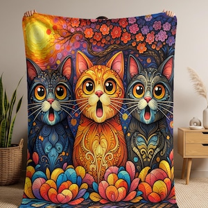 Può includere: Una coperta colorata con tre gatti stilizzati con occhi spalancati e bocche aperte. I gatti sono in tonalità di blu, arancione e grigio, su uno sfondo vibrante di fiori, un albero e un tramonto. Tessuto per la casa decorativo.