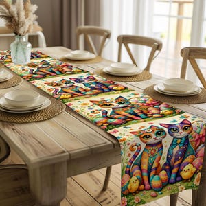 Può includere: Una colorata tovaglia a tema pasquale con illustrazioni di gatti stravaganti. I gatti sono raffigurati con strisce vibranti e dettagli floreali, circondati da uova di Pasqua e pulcini. La tovaglia è esposta su un tavolo di legno, aggiungendo un tocco di primavera.