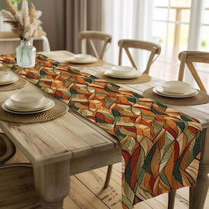 May include: A colorful table runner with an abstract leaf pattern in shades of orange, brown, green, and red. The runner is placed on a wooden dining table set with plates, placemats, and chairs. A vase with dried flowers is also visible.
