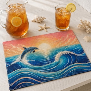Può includere: Una tovaglietta rettangolare con una scena dipinta di un delfino che salta fuori da onde blu verso un cielo al tramonto con nuvole arancioni e gialle. La tovaglietta è presentata con una caraffa di vetro e un bicchiere di tè freddo al limone.