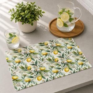 Può includere: Una tovaglietta rettangolare con un motivo botanico ripetuto di limoni gialli, foglie verdi e piccoli fiori bianchi su sfondo color crema. La tovaglietta è mostrata accanto a una caraffa di vetro con acqua e limone e una pianta in vaso.