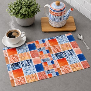 Può includere: Una tovaglietta rettangolare caratterizzata da un vivace design patchwork di quadrati blu, arancioni e rossi con vari motivi geometrici tra cui onde, punti e chevron, esposta con una teiera e una tazza di caffè.