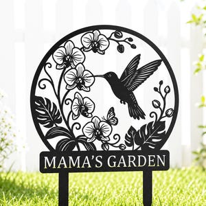 May include: A black metal garden stake featuring a circular frame with a cutout design of a hummingbird, orchid flowers, and tropical leaves. A rectangular plaque at the base displays the text MAMA'S GARDEN in bold, capitalised lettering.