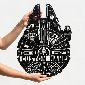 Może przedstawiać: Czarna, wycinana laserowo metalowa grafika ścienna w kształcie Sokoła Millennium. Projekt zawiera skomplikowane detale i tekst "CUSTOM NAME" w kolorze białym. Grafika jest trzymana przez dwie ręce na białym tle.