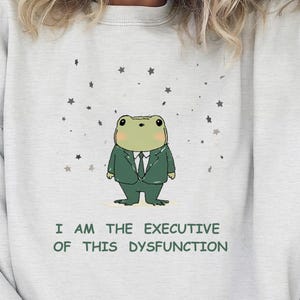 Könnte beinhalten: Hellgraues Sweatshirt mit einem Cartoon-Frosch im Anzug und Sternen. Der Text lautet "I AM THE EXECUTIVE OF THIS DYSFUNCTION". Ein lässiges, bequemes Oberteil.