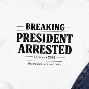 Puede incluir: Camiseta blanca con el texto "BREAKING PRESIDENT ARRESTED Caracas • 2026 History does not knock twice." La camiseta se combina con vaqueros azules y zapatillas blancas sobre una superficie de madera clara.