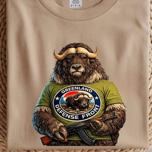 Puede incluir: Camiseta color arena con un gráfico de un buey almizclero que lleva una camiseta verde con el texto "Greenland Defense Front" y sostiene un rifle. La camiseta está doblada sobre una superficie circular tejida.