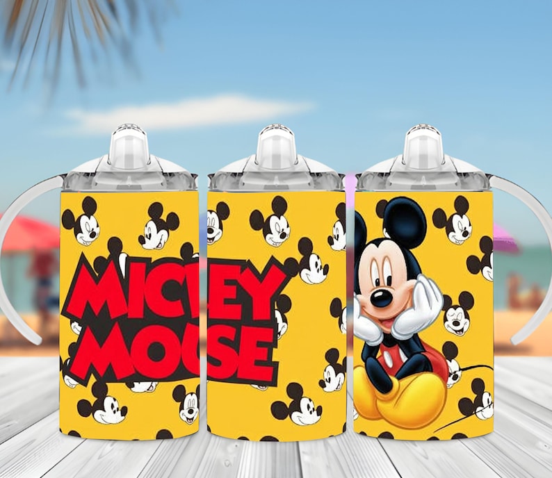 Puede incluir: Un vaso con boquilla amarillo con un patr&oacute;n repetitivo de cabezas de Mickey Mouse, tapa transparente y asas blancas. El texto MICKEY MOUSE aparece en letras rojas grandes en el lateral, junto a una ilustraci&oacute;n de Mickey Mouse apoyando su barbilla.