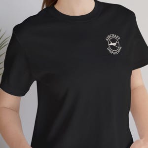 May include: A black short-sleeved t-shirt featuring a white circular logo on the upper left chest. The logo contains a silhouette of an airplane and the text AIRCRAFT DISPATCHER written in a circular border around the central graphic.