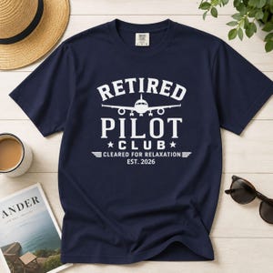 May include: A navy blue short-sleeve t-shirt featuring a white graphic print that reads "RETIRED PILOT CLUB" with an airplane icon, "CLEARED FOR RELAXATION", and "EST. 2026". The shirt has a crew neckline and a standard fit, displaying clear white text.