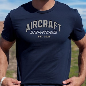May include: Navy blue t-shirt with the text "AIRCRAFT DISPATCHER EST. 2026" in a gray, arched font. The shirt has short sleeves and a crew neck. The design is centered on the chest.