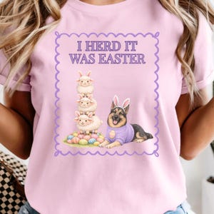 May include: Pink t-shirt with the text "I Herd It Was Easter" inside a purple scalloped border. The graphic features a stack of three sheep, a German Shepherd wearing bunny ears and a purple sweater, and a pile of Easter eggs.