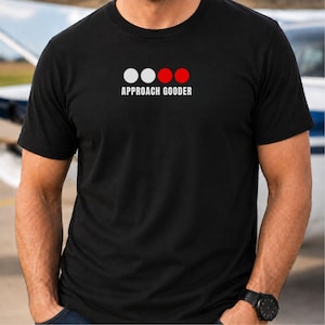 May include: A black short-sleeved t-shirt featuring a graphic design on the chest. The design consists of two white circles followed by two red circles, positioned above the white text that reads "APPROACH GOODER" in a clean, sans-serif font.