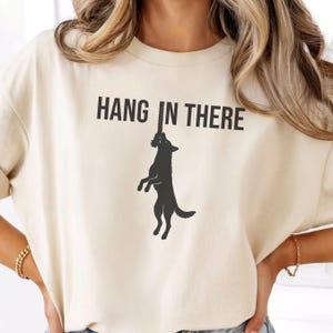 May include: A cream-colored t-shirt featuring a black graphic of a dog hanging from a rope. Above the dog, the text HANG IN THERE is printed in a bold, black, sans-serif font. The shirt has short sleeves and a crew neckline, providing a simple design.