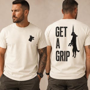 May include: Cream-colored t-shirt with a black dog head silhouette on the front and the words "GET A GRIP" with a dog climbing a rope on the back. The shirt is short-sleeved and worn by a person.