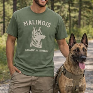 May include: An olive green t-shirt featuring the word "MALINOIS" and a dog illustration, with the text "GUARD & GUIDE" underneath. A person is wearing the shirt, standing next to a Belgian Malinois dog wearing a harness.