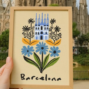 May include: A framed artwork featuring a stylized illustration of the Sagrada Familia in Barcelona, Spain. The artwork includes blue flowers, palm trees, and the word "Barcelona". The background shows the actual Sagrada Familia.
