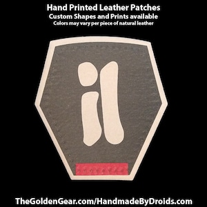 May include: A hand-painted leather patch in the shape of a hexagon with the letter "il" printed in white on a dark gray background. The patch has a red border and the text "TheGoldenGear.com/HandmadeByDroids.com" is printed below.