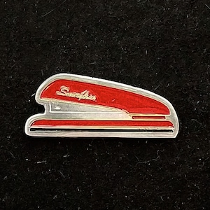 Op de afbeelding: Een rode en zilveren vintage nietmachine met het woord "Swingline" erop gedrukt. De nietmachine is een populair kantoorbenodigdheid.