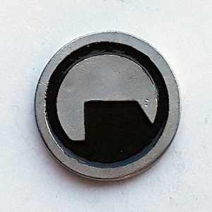 Puede incluir: Un pin circular de color plateado con un símbolo en blanco y negro. El símbolo es un círculo con una forma negra en el interior que se asemeja a un edificio o una puerta.