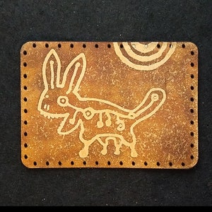 May include: A brown leather patch with a white hand-painted design of a stylized rabbit. The patch has a stitched border and is available in custom shapes and prints.