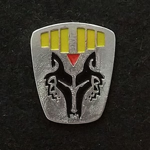 May include: A silver-colored metal pin with a black wolf head design. The wolf's eyes are red, and there are five yellow bars above the wolf's head.