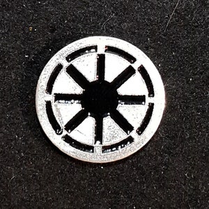 Può includere: Uno spillo color argento con un simbolo bianco e nero di un cerchio con otto raggi che irradiano dal centro.