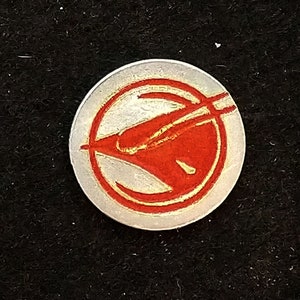 Può includere: Uno spillo in metallo argentato con un designo rosso e oro. Il design presenta un uccello stilizzato in volo. Lo spillo è circa delle dimensioni di una moneta da 20 centesimi di euro.