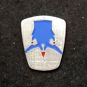 May include: A silver-colored metal pin with a blue and red enamel design. The pin is shaped like a shield and features a stylized bird with outstretched wings. The bird is blue with red accents on the wings and beak.