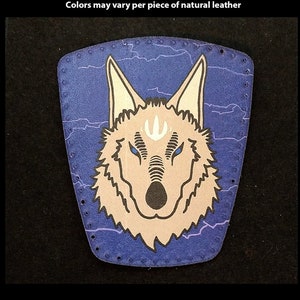以下が含まれることがあります： 青い背景に青い目をした狼の頭が描かれた、手描きの革のパッチ。パッチはステッチされた縁取りがされており、カスタムの形状とプリントが可能です。