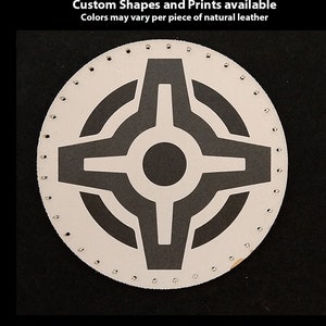 May include: A white leather patch with a black geometric design. The patch has a circle in the centre with four arms extending out. The patch is surrounded by a circle of small holes.