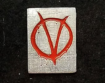 May include: A silver metal pin with a red V for Vendetta symbol. The pin is about the size of a US quarter.