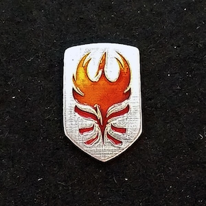 May include: A silver shield-shaped pin with a red and orange phoenix design. The phoenix is stylized with a flame-like design and is set against a textured silver background.