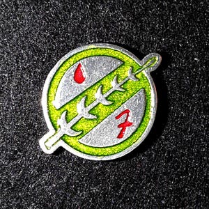 May include: A silver pin with a green and white design. The design features a stylized tree with a red drop of blood at the top. The number "7" is visible on the pin.