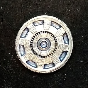 May include: A silver-colored metal coin with a circular design featuring a concentric circle pattern and a central circle. The design resembles a stylized version of the Iron Man arc reactor.