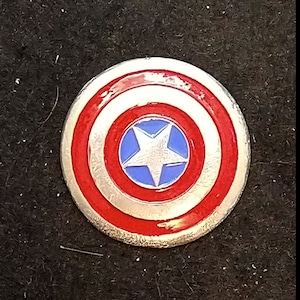 May include: A silver-colored Captain America shield pin with a red, white, and blue design. The shield is about the size of a US quarter.