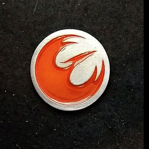 Puede incluir: Un pin de color plateado con un símbolo rojo de la Alianza Rebelde. El símbolo es un pájaro estilizado blanco en vuelo con tres alas. El pin es aproximadamente del tamaño de una moneda de 25 centavos de dólar estadounidense.